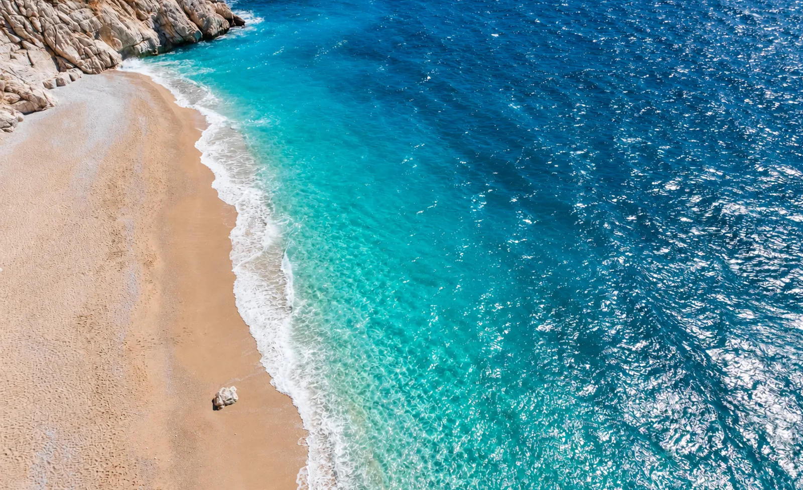 Sunlit turquoise shallows and white surf along Kaputaş Beach, viewed from above